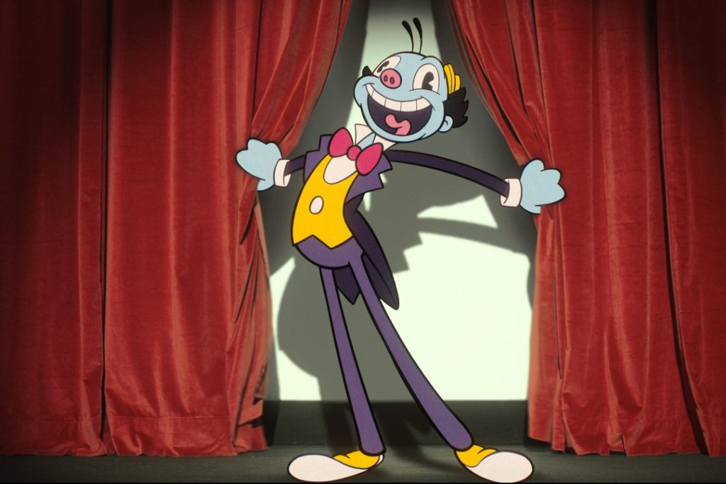 Cartoon character in a tuxedo and bow tie, smiling broadly while standing between red curtains on a stage.