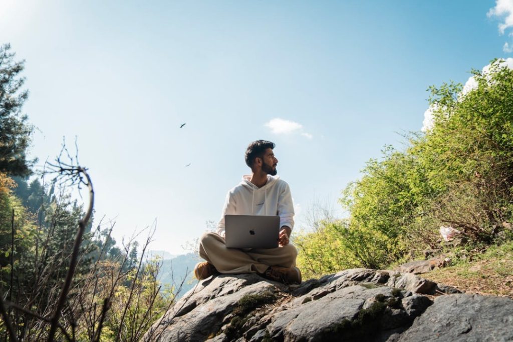 Man sitting on a rock outdoors, using a laptop. He is surrounded by greenery and a clear sky.
