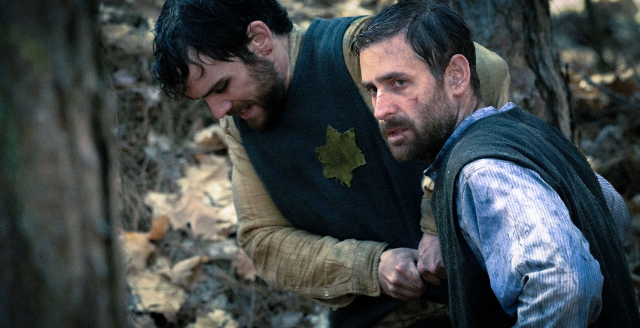 Two men are in a forest, one helping the other to his feet. The man on the right looks forward with a serious expression. They are dressed in rustic clothing among fallen leaves.