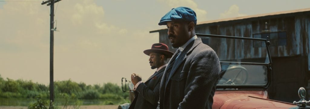 Two men in vintage suits and hats stand by a red convertible car near a rustic building on a sunny day.