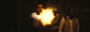 A man in a vest fires a gun indoors, producing a bright muzzle flash. Another man stands beside him, looking in the same direction. The setting appears dimly lit.