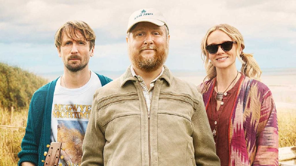 Three adults stand outdoors in a field, looking at the camera; one holds a guitar, another wears a cap, and another wears sunglasses.