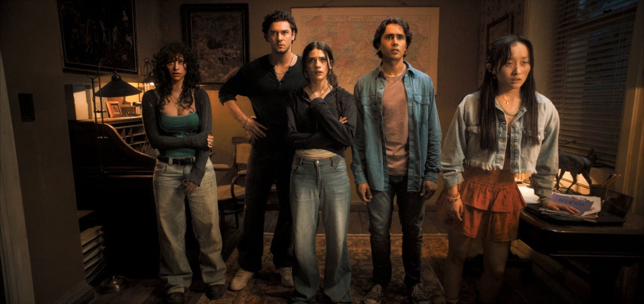 Five people stand indoors in a dimly lit room, all facing forward with serious expressions, surrounded by scattered papers and furniture.