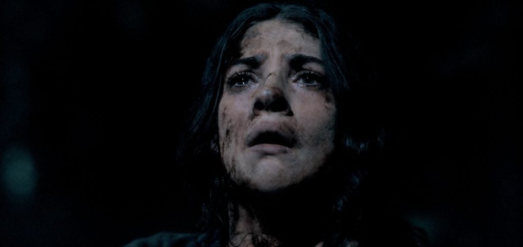 A woman with dirt on her face looks upward with a distressed expression against a dark background.