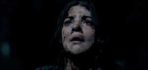 A woman with dirt on her face looks upward with a distressed expression against a dark background.