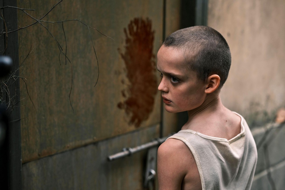 A young person with a shaved head and white tank top stands near a locked metal door with a reddish handprint stain on it.