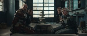 Four people in tactical gear sit tied to chairs in a dimly lit, industrial room, looking toward the camera.