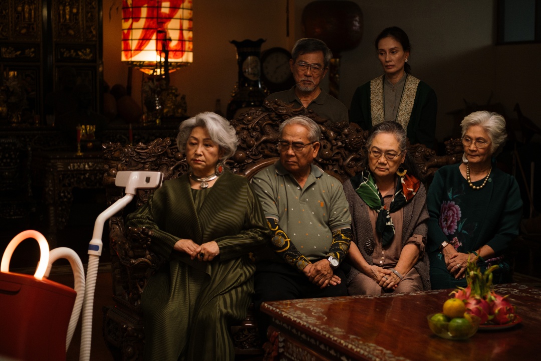 Six elderly people sit together in a dimly lit room, facing a robot with a screen. The atmosphere appears serious and attentive.