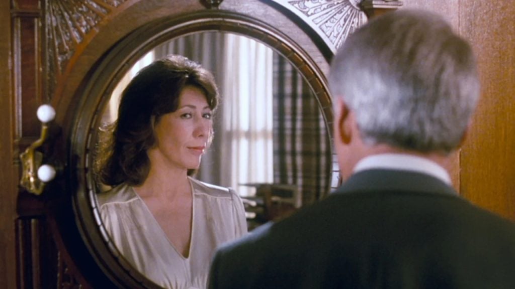 A woman looks at her reflection in an ornate round mirror, while a man with gray hair stands in front of her, facing the mirror.
