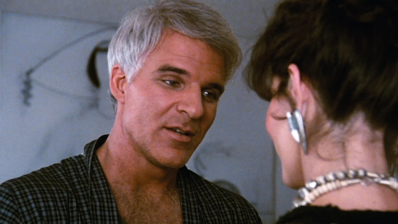 A man with gray hair and a plaid robe speaks to a woman wearing large earrings and a pearl necklace; both are indoors.
