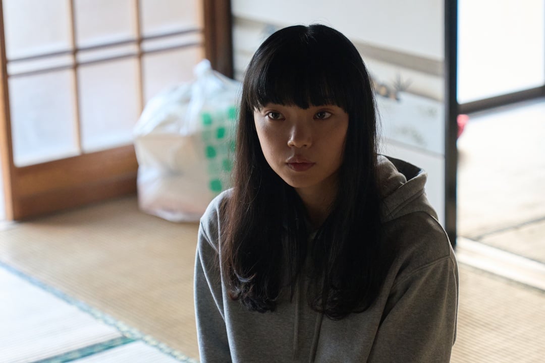 A young woman with long black hair and bangs sits indoors wearing a gray hoodie, with a paper sliding door and a plastic bag in the background.