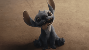 A small, blue creature with large ears and big eyes sits on the ground, looking up with its mouth open and tongue out.