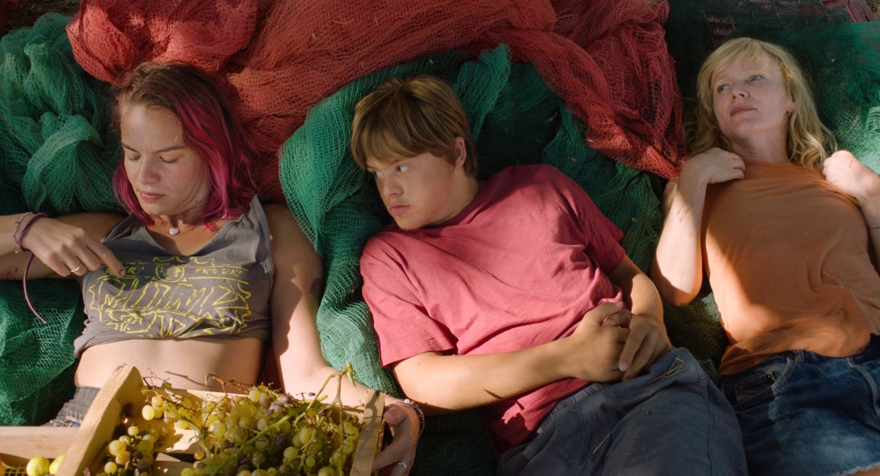 Three people lie on colored nets outdoors, with one holding a tray of grapes. They appear relaxed and are dressed in casual clothing.