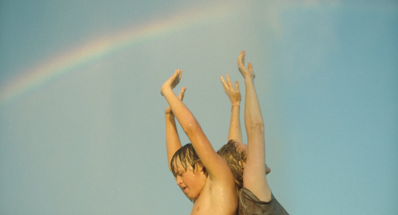 Two children with wet hair raise their arms against a blue sky with a faint rainbow in the background.
