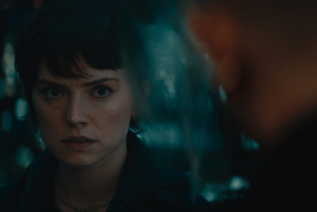 A person with short dark hair and a serious expression looks intently at someone who is out of focus in the foreground.