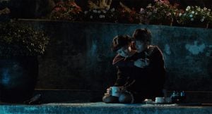 Two people sit closely together on a blanket at night, with one person comforting the other; tea and flowers are visible nearby.