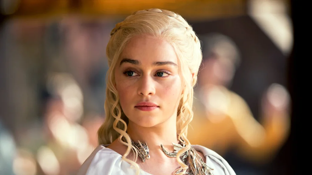 A woman with long, braided blonde hair and a silver necklace looks to the side, wearing a white outfit in a softly lit indoor setting.