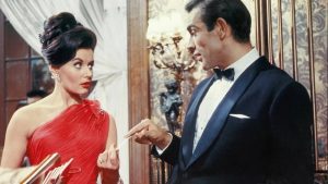 A woman in a red dress and a man in a black tuxedo are talking indoors; the man gestures with his hand while the woman looks at him with a surprised expression.