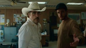 Two men stand in an office; one wears a white cowboy hat and glasses, the other wears a dark cap and gestures forward. The room has desks, papers, and maps on the walls.