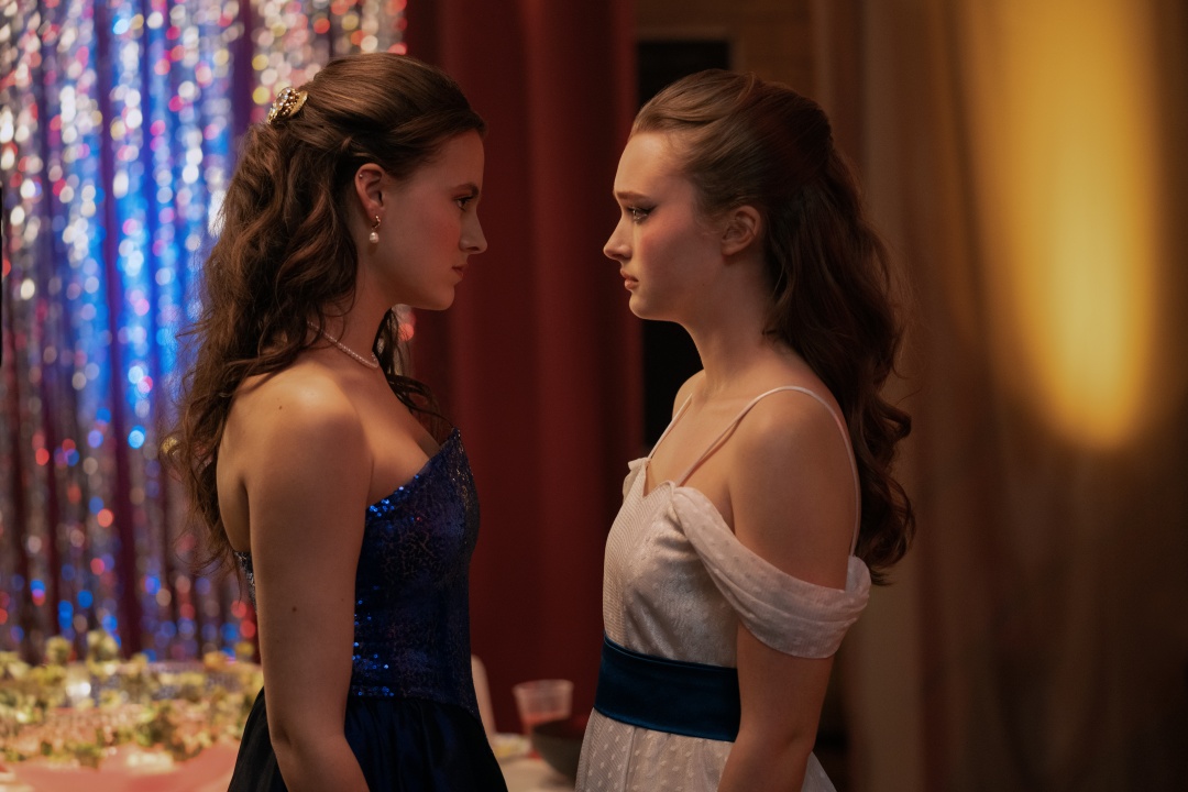Two young women in formal dresses stand face-to-face at an event with a decorated backdrop and a table with drinks and food in the background.