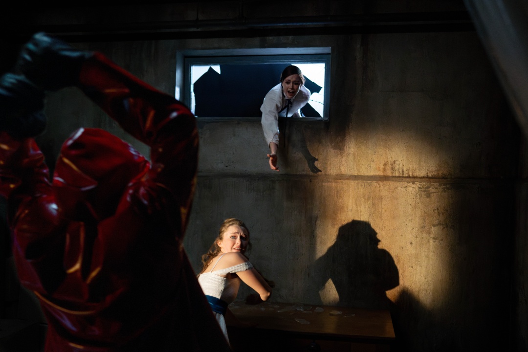 A woman in a white shirt reaches through a high window toward a frightened woman seated below as a person in a red hooded outfit raises an object menacingly.