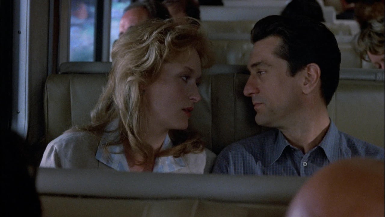 A woman and a man sit closely together on a bus, looking into each other's eyes, with other passengers seated around them.