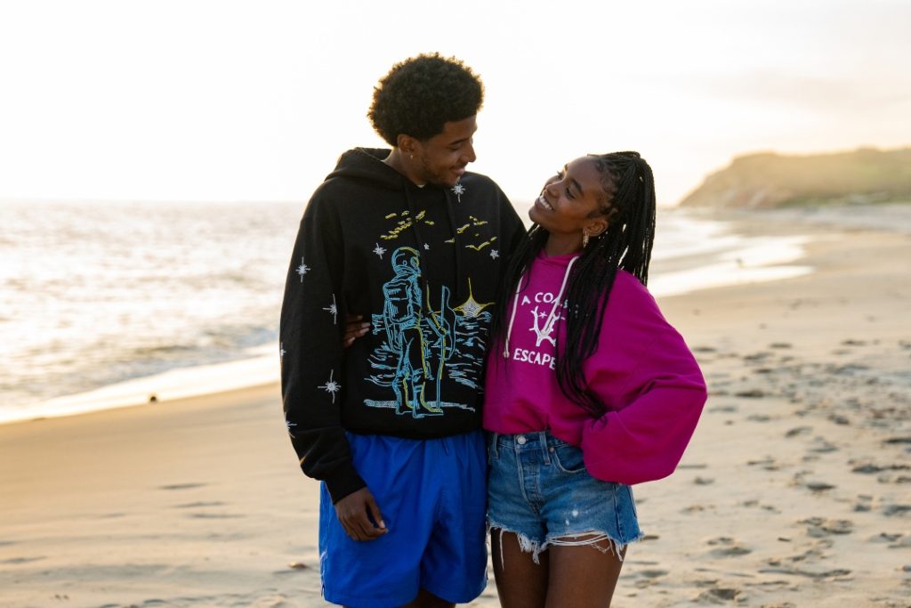 Two people stand close together on a sandy beach at sunset, smiling at each other. One wears a black hoodie and blue shorts, the other a pink sweatshirt and denim shorts.