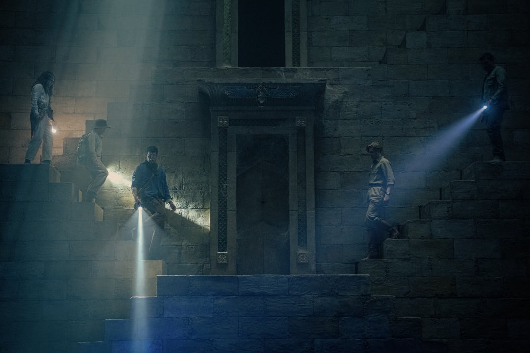 Five people on ancient stone steps shine flashlights at a large, ornate doorway set into a wall illuminated by beams of light.