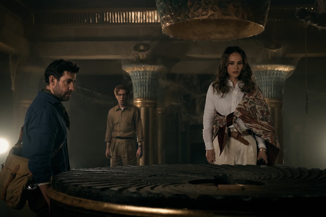 Three people stand around a large, circular ancient artifact in an ornate, dimly lit room with Egyptian-style columns.