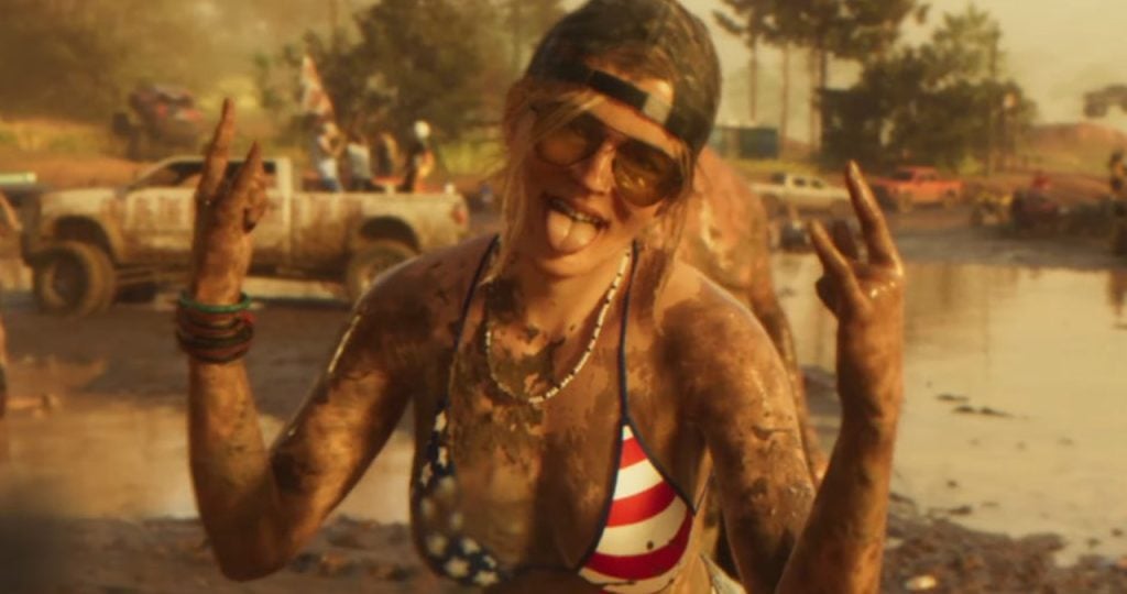 A woman covered in mud wearing sunglasses, a backward cap, and a stars-and-stripes bikini top makes hand gestures and sticks out her tongue at an outdoor muddy event.