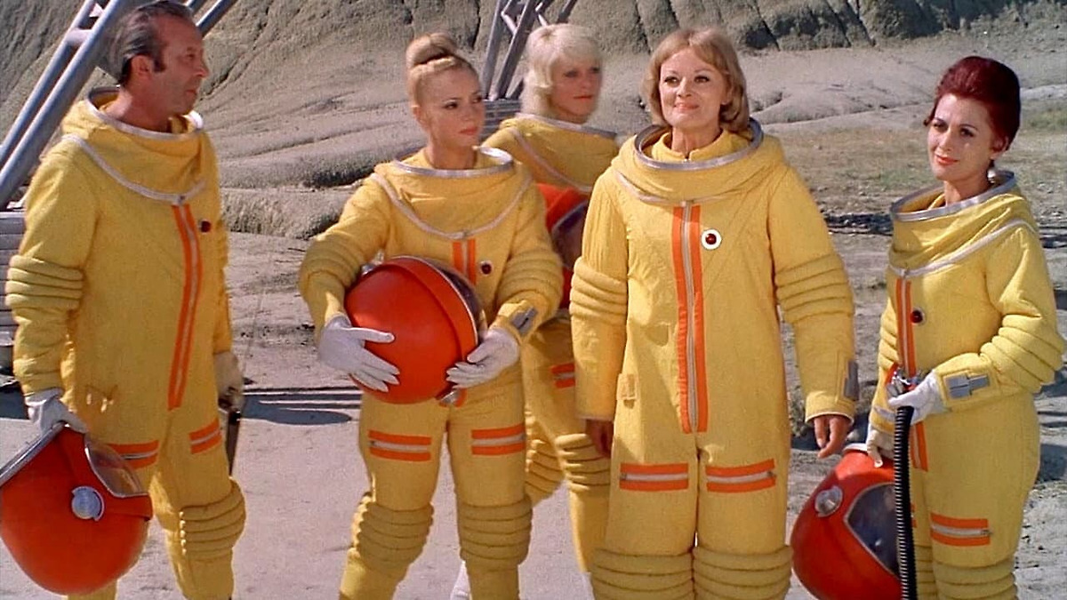 Five people in yellow space suits stand outdoors on rocky terrain, holding orange helmets and equipment, with a metal structure visible on the side.