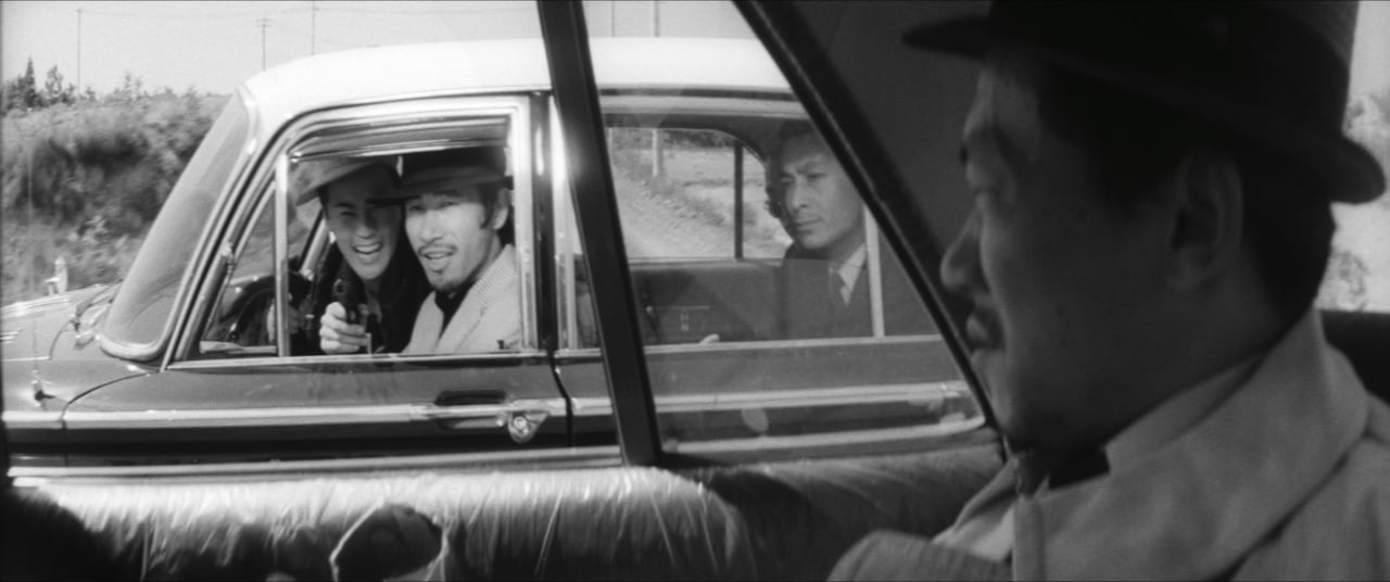 Two men in a car point a gun through the window at a man sitting in the back seat of another car, with a driver visible in the foreground.
