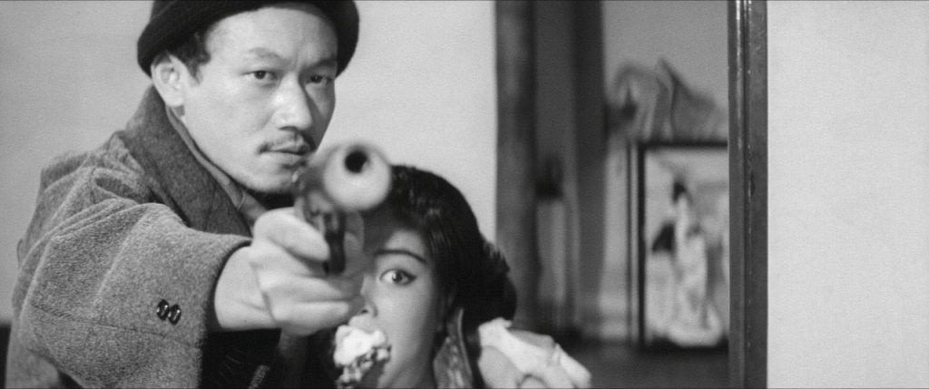 A man in a coat and hat points a pistol toward the camera while a woman sits closely behind him, partially obscured and looking in the same direction.