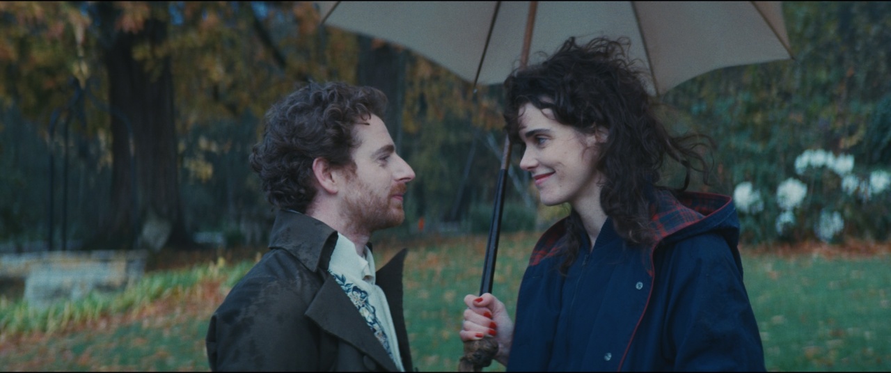 A man and a woman stand close together outdoors under an umbrella, facing each other and smiling, with blurred trees and greenery in the background.