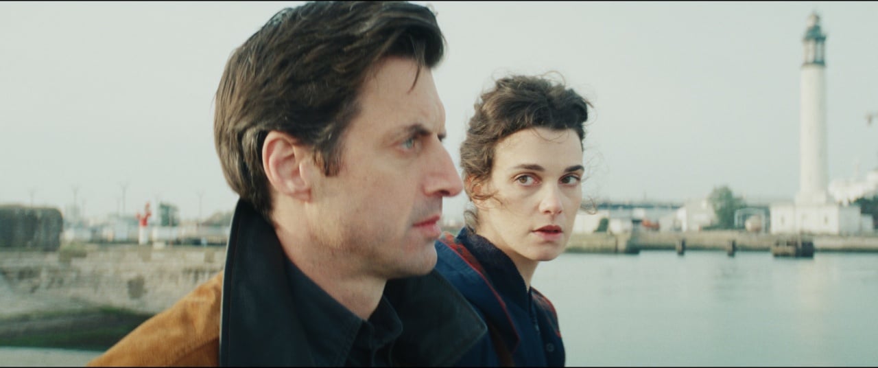 A man and a woman stand near a body of water with a lighthouse in the background. The woman looks toward the camera while the man looks ahead.