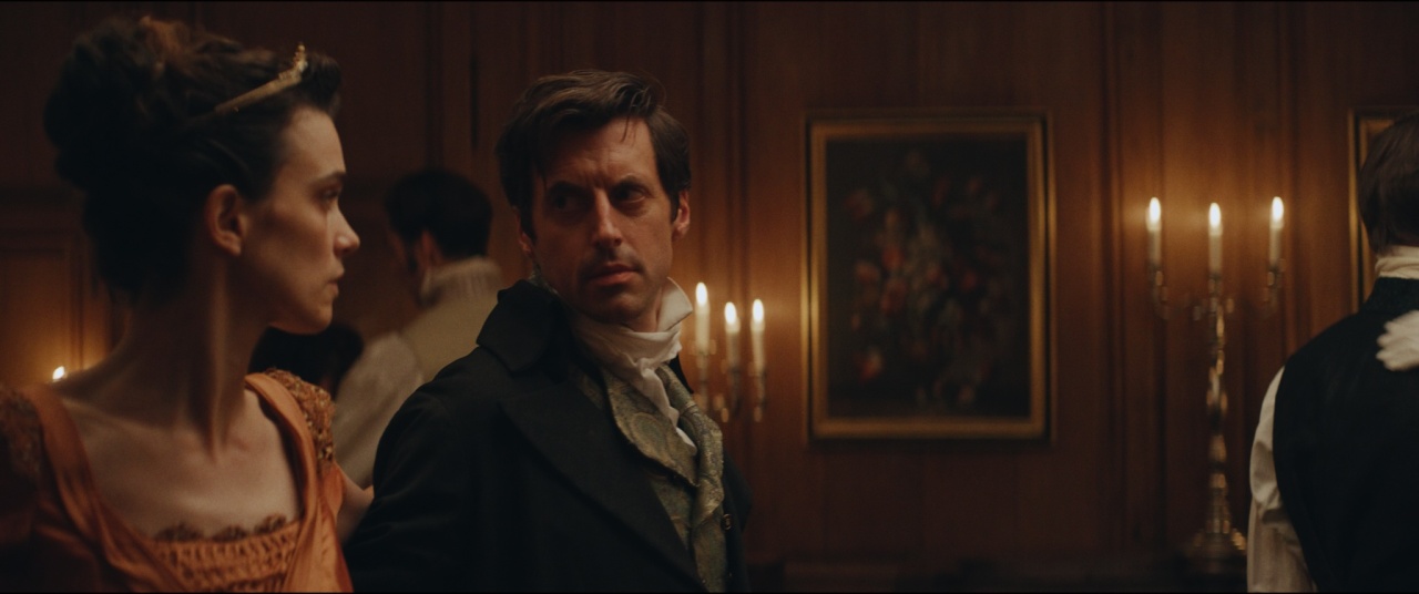 Two people in historical attire stand in a dimly lit room with wood-paneled walls, candles, and framed artwork in the background.