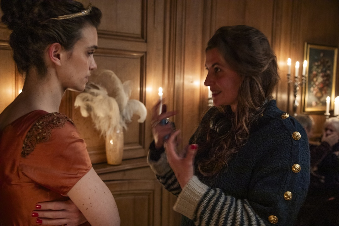 Two women are indoors having a serious conversation; one wears an orange dress, the other a dark coat with gold buttons, with candles and a feather arrangement in the background.
