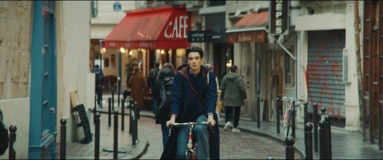 A young man rides a bicycle down a narrow city street lined with shops, including a red-awning café, while pedestrians walk in the background.