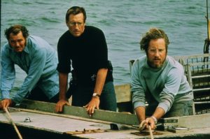 Three men lean over the edge of a boat, holding ropes and looking intently at something off frame with water in the background.