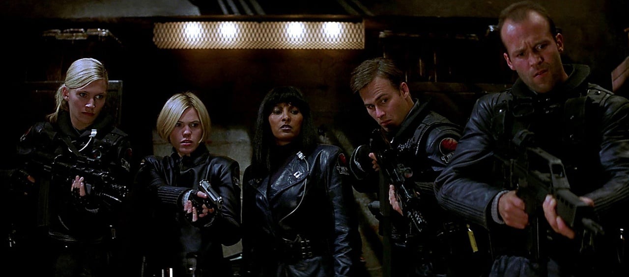 Five people in black tactical gear stand close together in a dimly lit room, four aiming guns forward while one stands in the center watching intently.