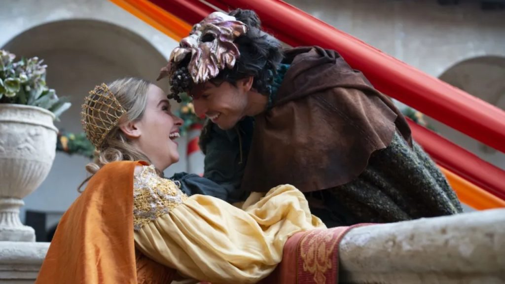 Two people in historical costumes, one wearing a mask and cloak, lean close to each other and smile in an outdoor setting decorated with red and orange fabric.