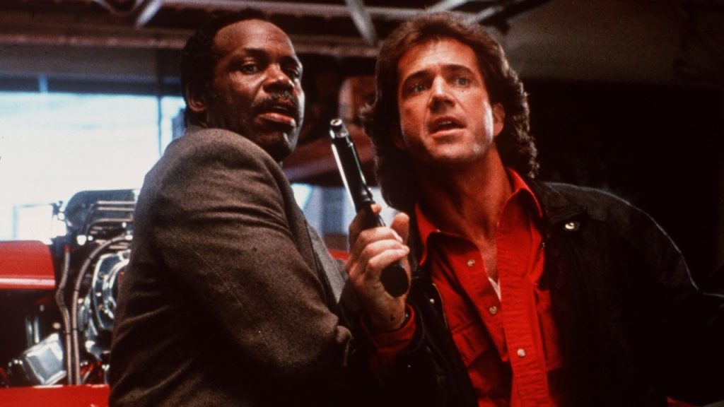 Two men stand close together indoors; one holds a gun while the other looks in the same direction, appearing tense.