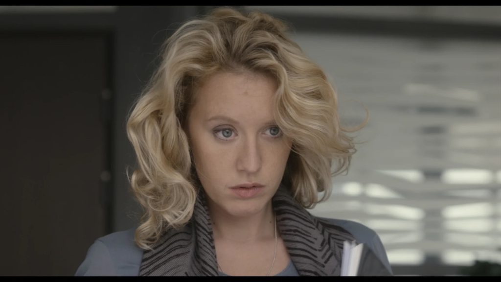 A woman with wavy blonde hair and a gray patterned scarf looks ahead with a serious expression, holding papers in her arm.