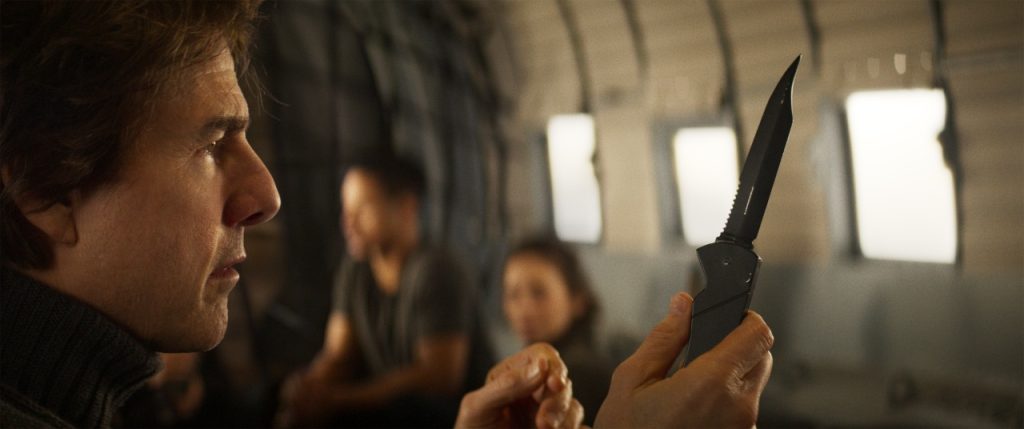 A man inside an aircraft holds and examines a black folding knife, with two other people sitting blurred in the background.