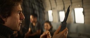 A man inside an aircraft holds and examines a black folding knife, with two other people sitting blurred in the background.