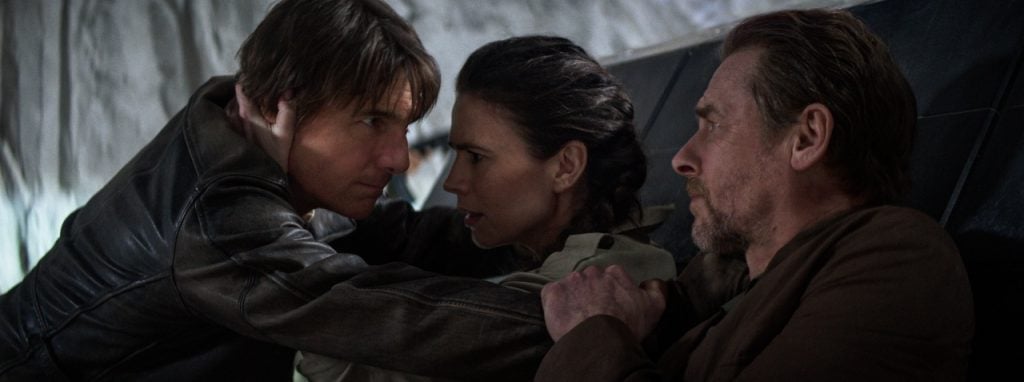 Three people sit closely together in a tense moment, with one man facing a woman who has her arms on his shoulders, while another man looks on from the side.