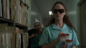 Cult Horror Favorite ‘May’ Heads To Blu-Ray Very Soon As A Part Of The Vestron Video Collector’s Series A woman in medical scrubs and dark glasses holds blood sample vials in a hallway lined with files; a man in scrubs sits in the background.