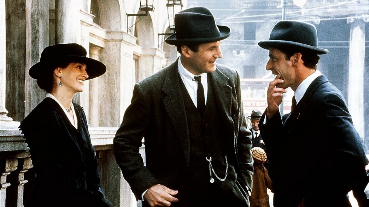 Three people in 1940s-style formal clothing and hats stand outdoors, engaged in conversation and smiling, with old stone buildings in the background.