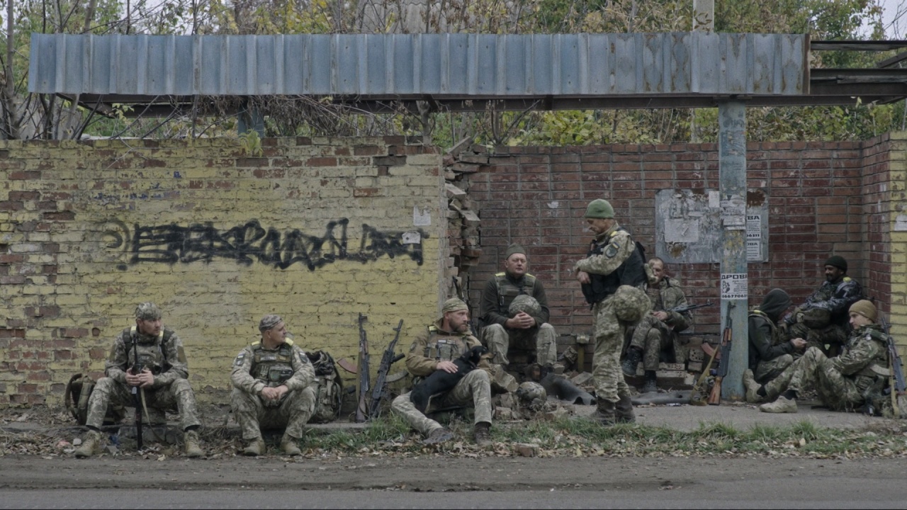 A group of soldiers in camouflage uniforms sit and rest with their gear against a dilapidated brick wall and metal shelter.