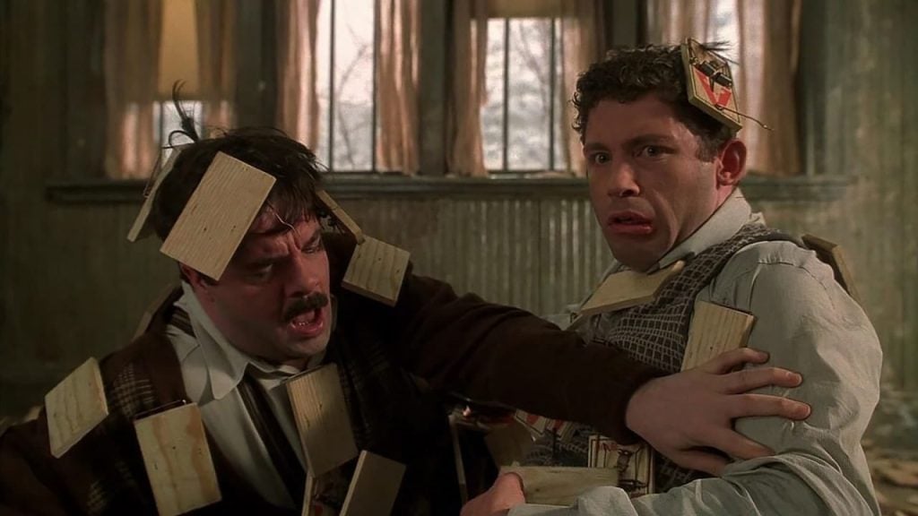 Two men sit on the floor, covered in wooden boards with shocked expressions, inside a room with peeling green walls and sunlight coming through the windows.